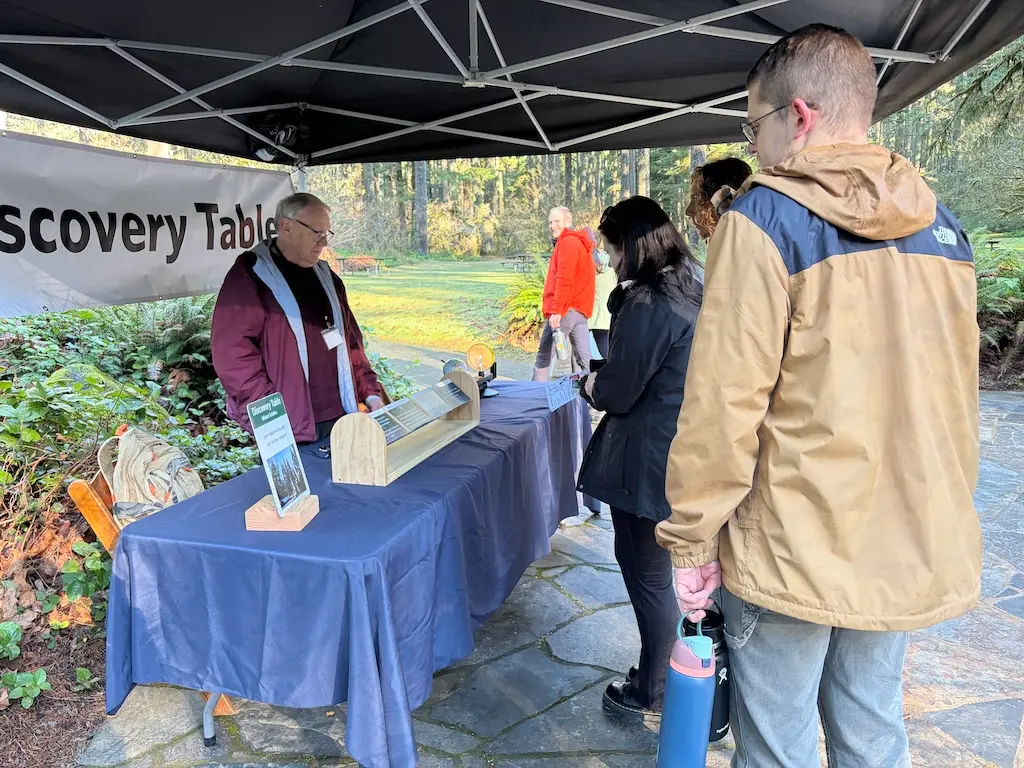 Park ranger hosts outdoor Discovery Table booth under tent, greeting visitors in forested park setting with stone patio.