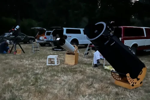 Amateur astronomers setting up Dobsonian and refractor telescopes on a grassy field at a dusk star party gathering.