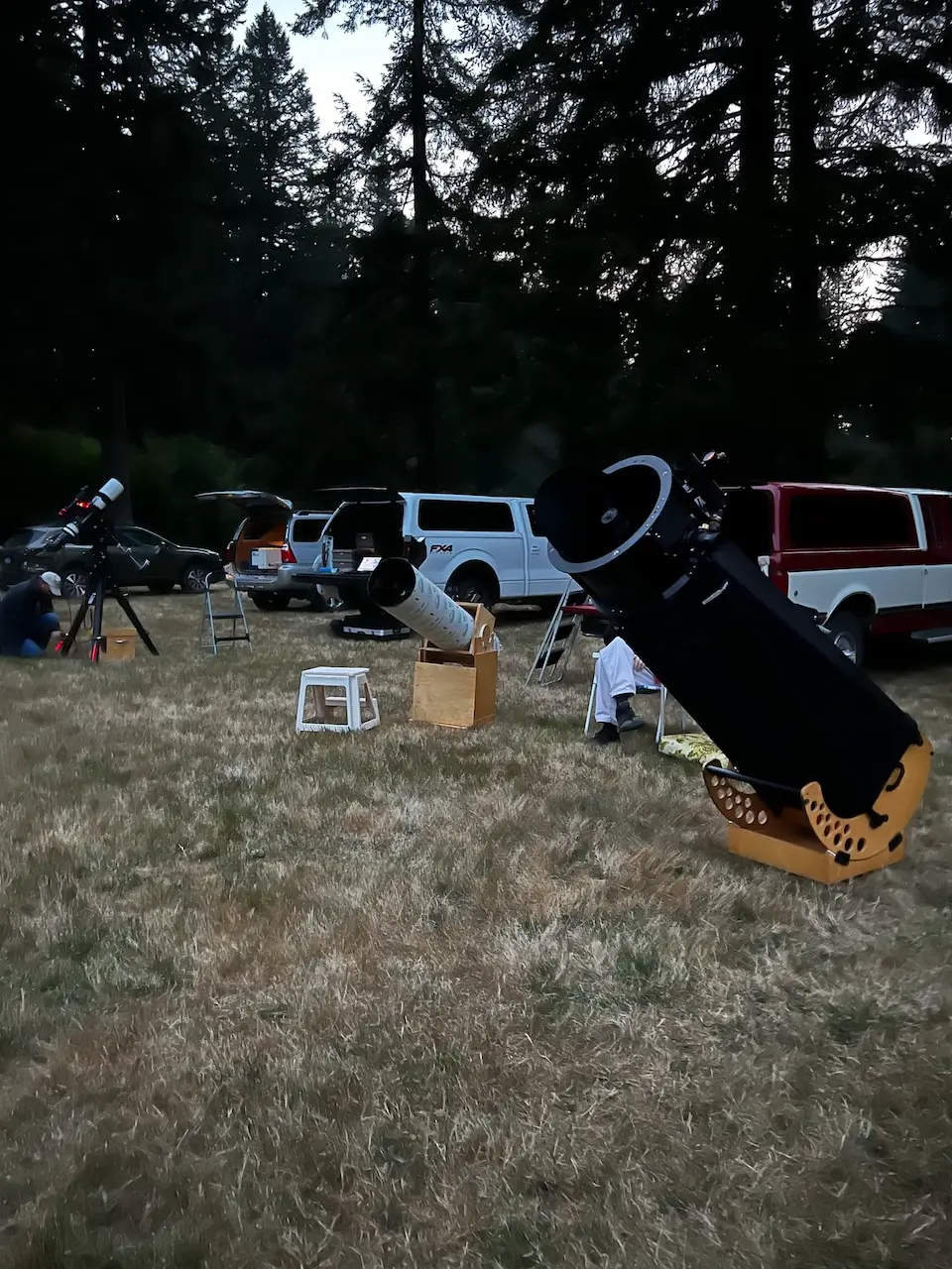 Amateur astronomers setting up Dobsonian and refractor telescopes on a grassy field at a dusk star party gathering.