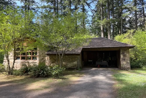 stone kitchen shelter Rustic stone kitchen shelter with cedar shingle roof nestled among tall evergreen trees in a sunlit forest park setting.
